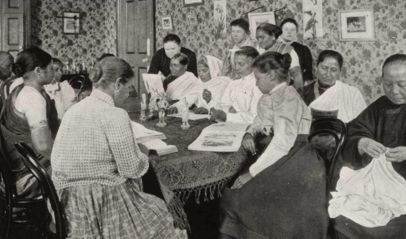 Interior scene with a large group of women seated around a table, many of them looking at documents.